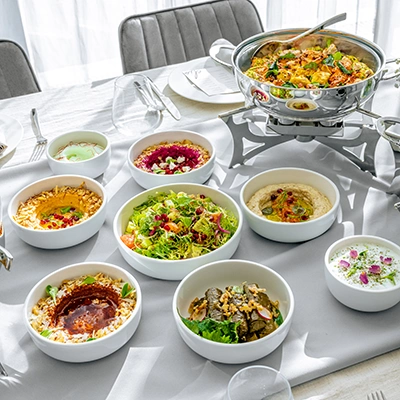 A dining table set with mezze dishes in white bowls, including hummus, salad and stuffed vine leaves, beside a serving pan