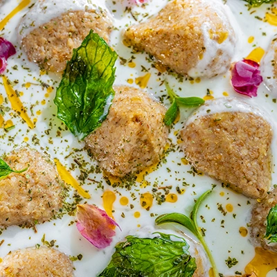 Close-up of kibbeh in yoghurt sauce, garnished with crispy basil, dried mint, rose petals and a drizzle of oil