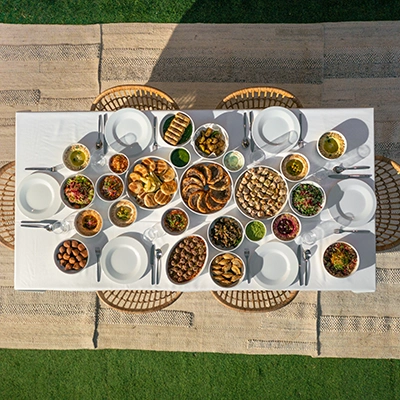 Overhead view of a long outdoor table filled with mezze and main dishes, set on a woven rug over grass